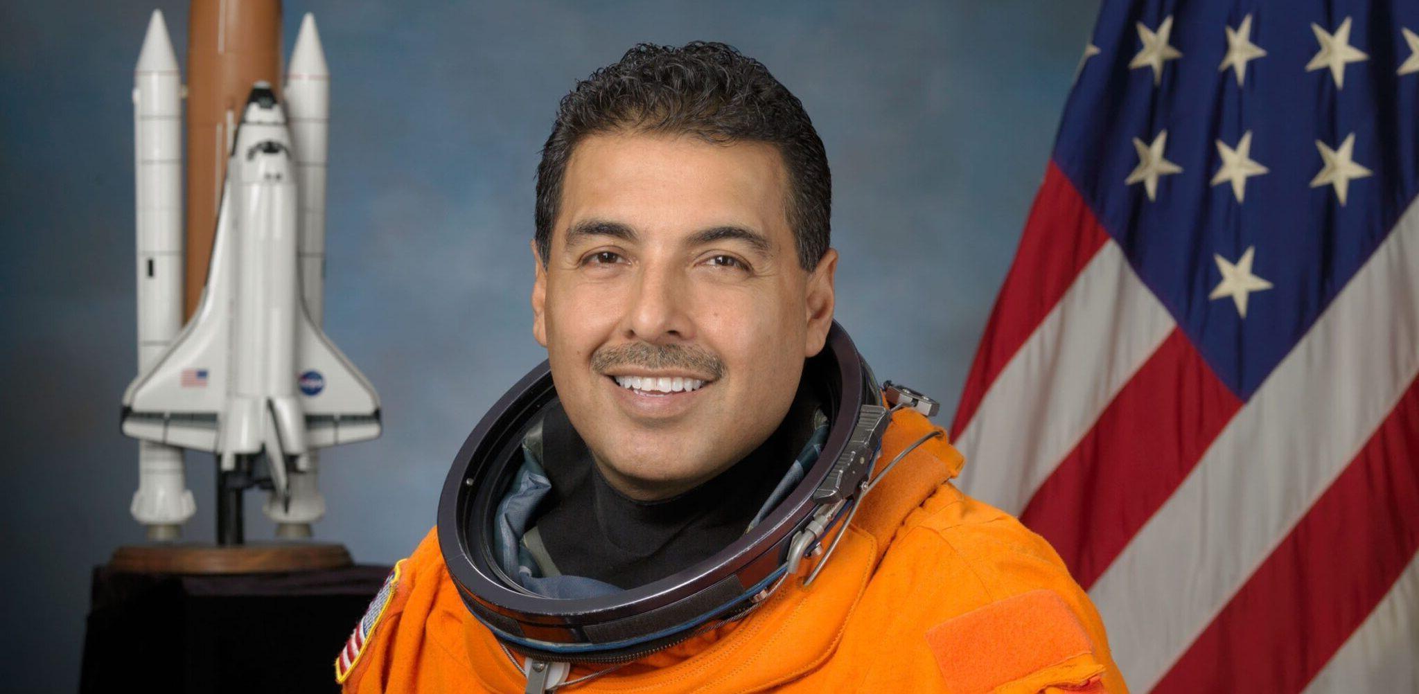 Astronaut Jose M. Hernandez, mission specialist in Orange space suit, American flag and Space shuttle model in the background